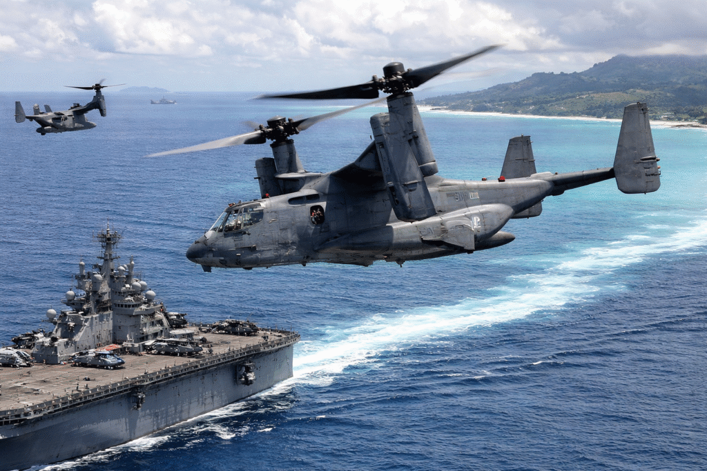 Two U.S. military Osprey tiltrotor aircraft fly above a U.S. Navy amphibious assault ship in the Caribbean Sea, with additional naval vessels and a tropical coastline visible in the background.