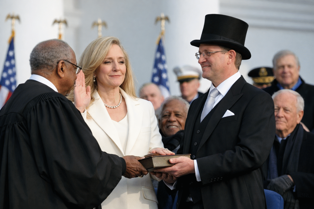 Abigail Spanberger raises her right hand while taking the oath of office as Virginia governor on the South Portico of the Virginia State Capitol in Richmond, standing beside her husband holding a Bible, with officials and American flags in the background.