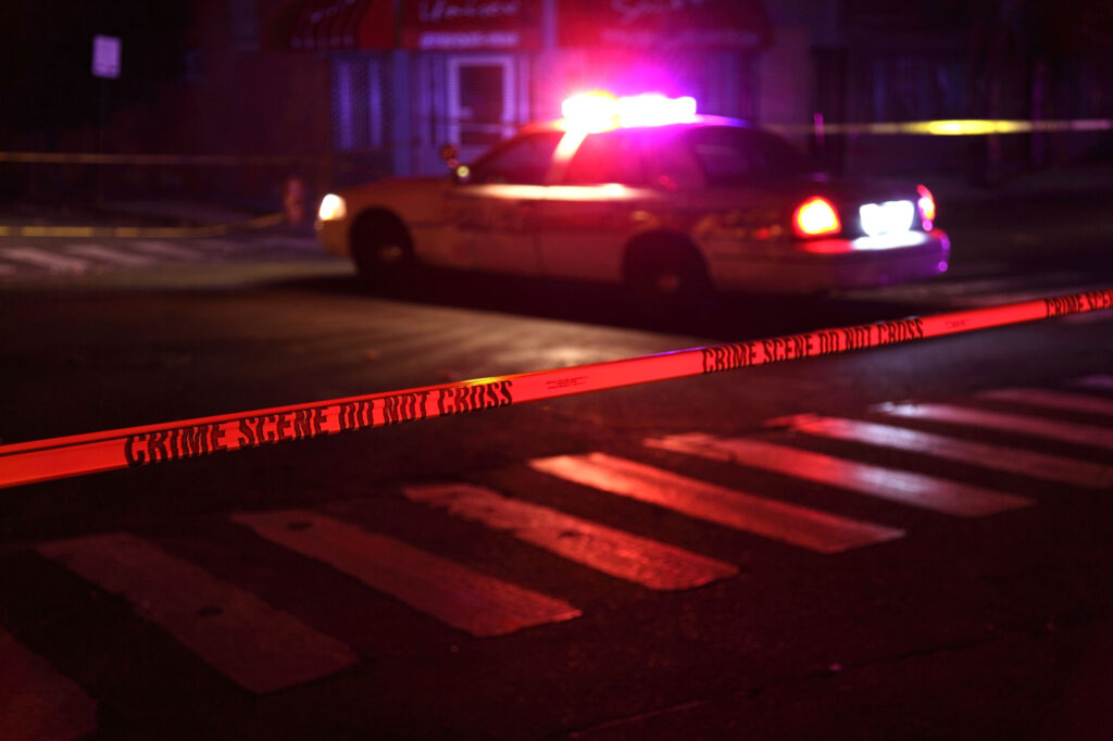 Dark intelligence briefing room with classified documents on a desk, surveillance screens glowing in the background, and a cordoned-off crime scene at night, symbolizing covert intelligence activity, surveillance networks, and a compromised investigation.