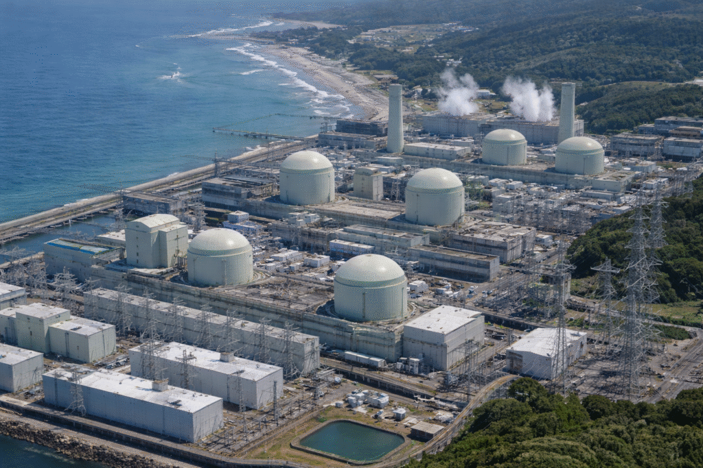 Aerial view of the Kashiwazaki-Kariwa nuclear power plant along Japan’s coastline, showing multiple reactor domes, industrial buildings, transmission lines, and the Pacific Ocean in the background.