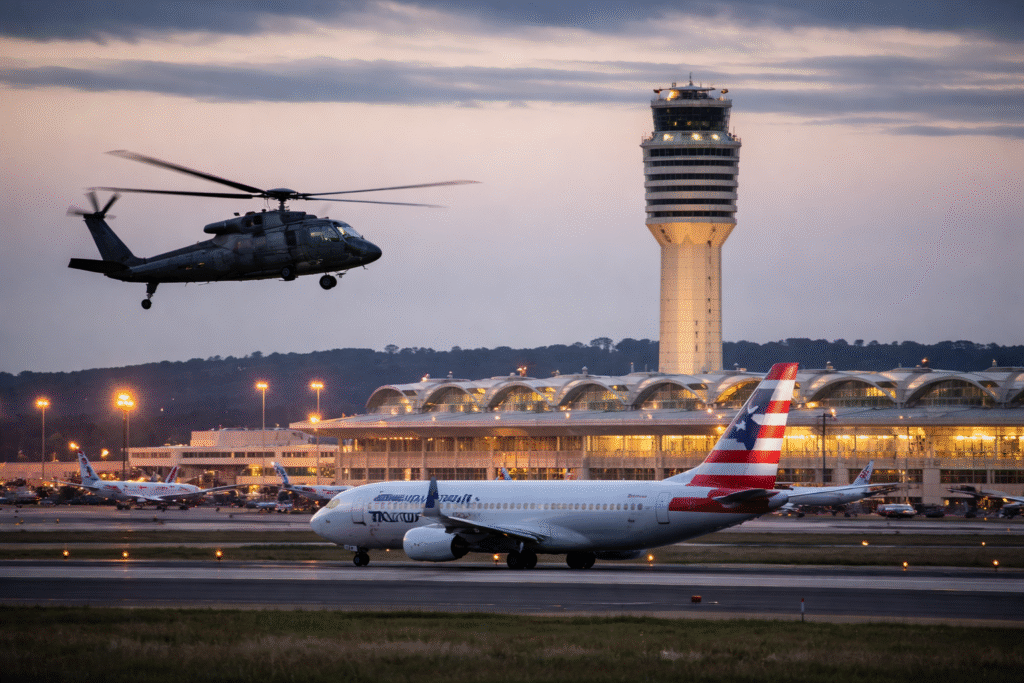 American Airlines commercial jet taxiing at Reagan National Airport with a U.S. Army Black Hawk helicopter flying nearby and the airport control tower visible in the background at dusk.