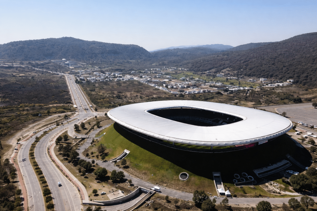 Aerial view of a large oval stadium with a white roof set into a grassy hillside, surrounded by winding roads and a small town in a mountainous region under clear daylight skies.
