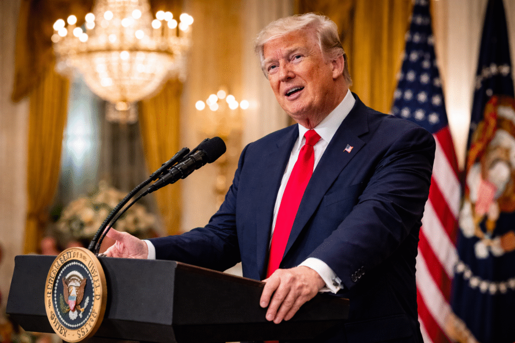 Donald Trump speaking at a podium in a formal White House setting, wearing a navy suit and red tie, with American flags and chandeliers in the background.