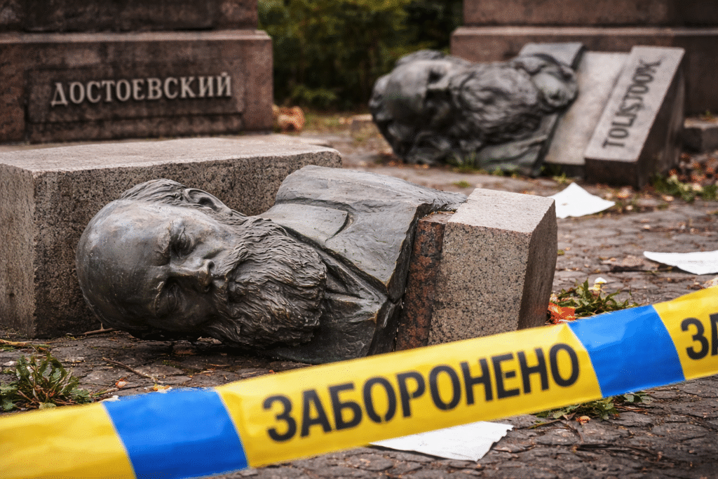 Toppled bronze busts of Fyodor Dostoevsky and Leo Tolstoy lie on the ground beside empty stone pedestals, partially cordoned off with blue and yellow Ukrainian police tape in an outdoor public space.