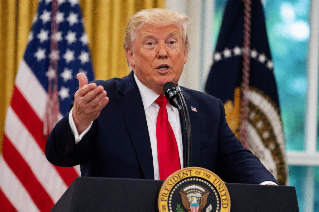 Donald Trump speaking at a podium with a raised hand gesture, wearing a dark suit and red tie, with the US flag and presidential flag behind him.