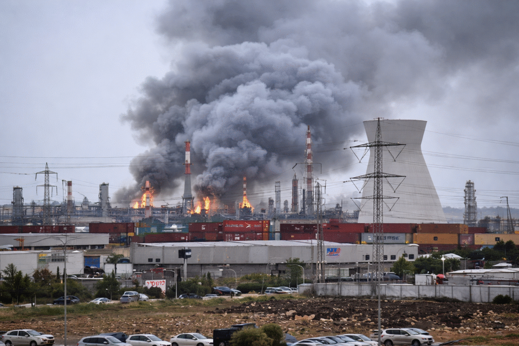 Industrial refinery complex with cooling towers and smokestacks emitting thick gray smoke and visible flames, with shipping containers, power lines, and vehicles in the foreground.