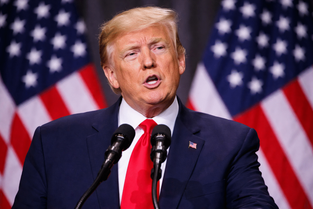 Donald Trump speaking at a podium with microphones, wearing a dark suit and red tie, with blurred American flags in the background.