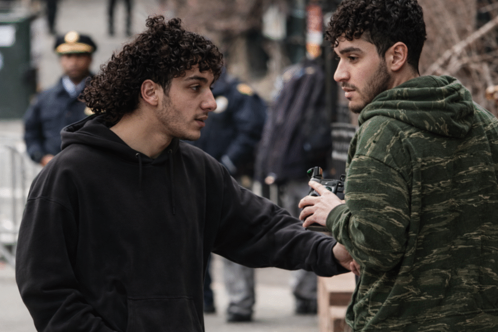 Two young men standing on a city sidewalk speaking with each other while police officers and barricades are visible in the background.