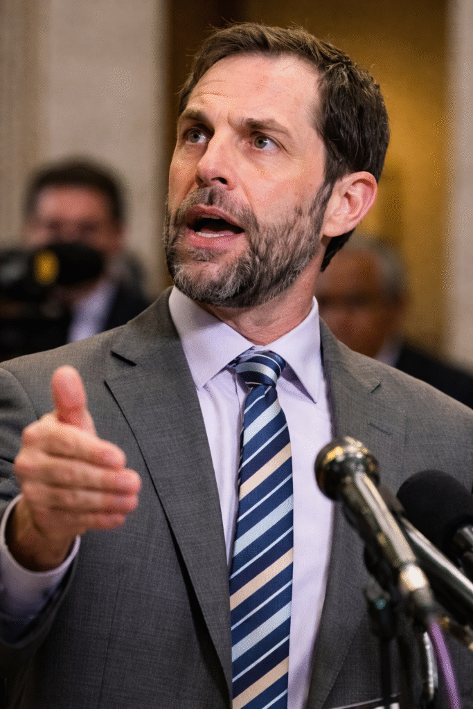 Close-up of a man with short dark hair and a trimmed beard speaking at a press conference, wearing a gray suit and striped tie, gesturing with his hand beside microphones.