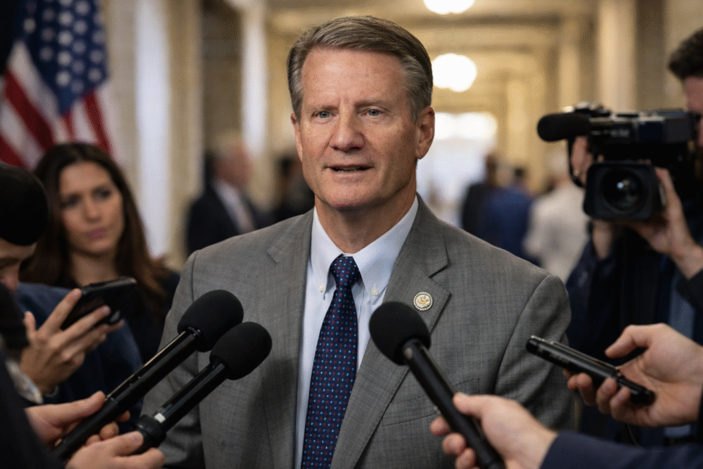 Rep. Tim Burchett speaking to reporters in a Capitol hallway with multiple microphones pointed toward him and an American flag in the background.
