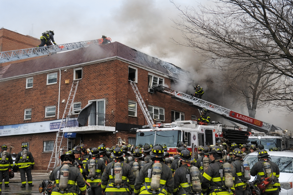 Firefighters in full protective gear using ladders and a fire truck to battle a heavy blaze at a multi-story brick building, with thick smoke rising from the roof.