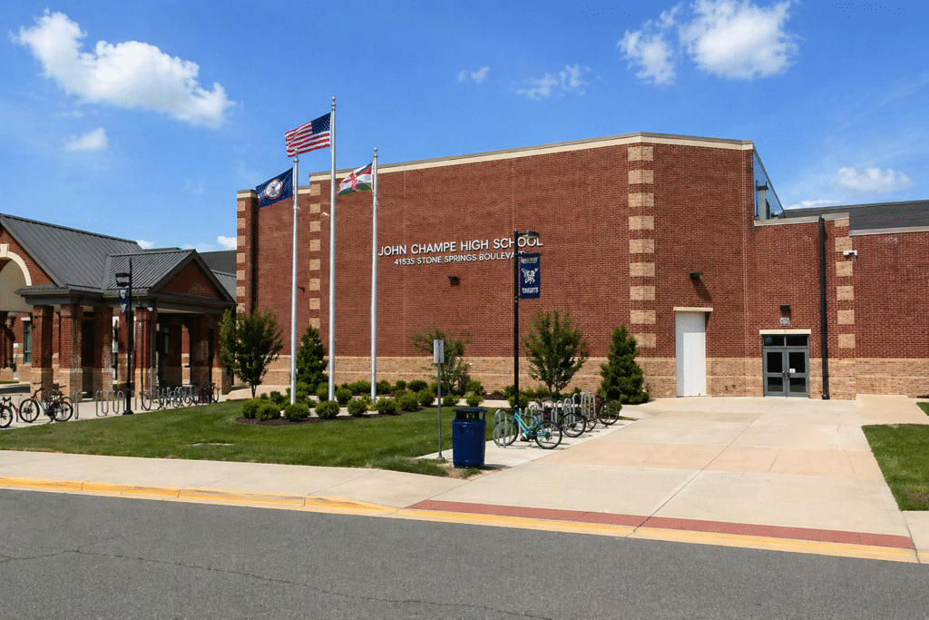 Exterior of John Champe High School in Stone Ridge, Virginia, showing a red-brick building, flagpoles with U.S. and state flags, and a landscaped entrance area on a clear day.