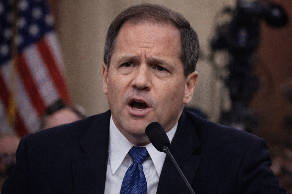 Man in a suit speaking into a microphone during a formal hearing, with an American flag visible in the background.
