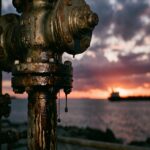 Close-up of an oil pipeline valve dripping crude oil at sunset with a cargo ship blurred in the background over the ocean.