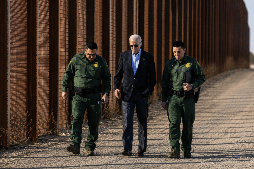 President Joe Biden walks alongside two Border Patrol agents near a tall border fence in a desert area during an official border visit.