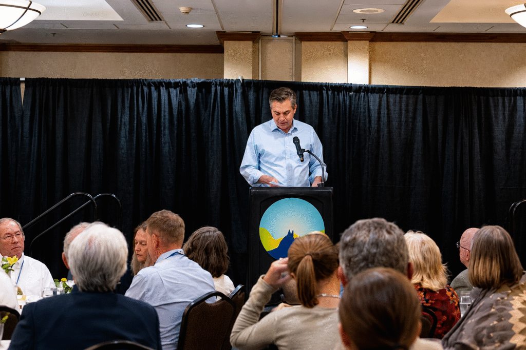 Jim Acosta speaking at a podium during a Montana Democratic event while attendees sit at banquet tables in a conference room.