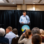 Jim Acosta speaking at a podium during a Montana Democratic event while attendees sit at banquet tables in a conference room.