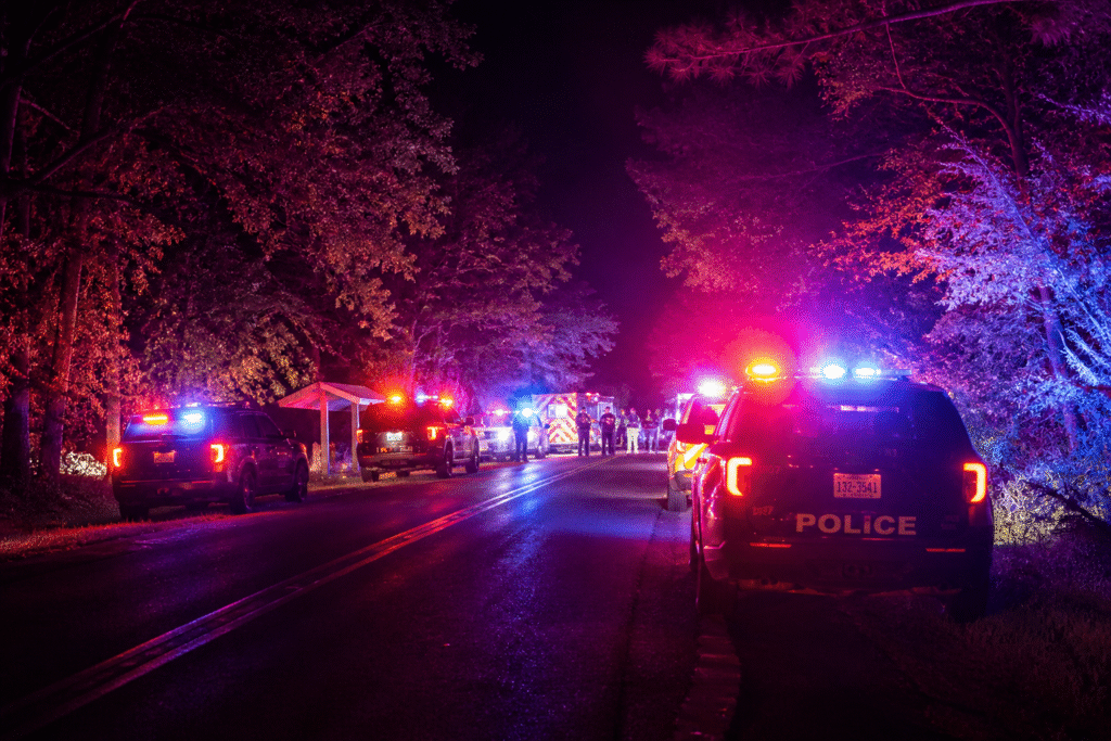 Police vehicles with flashing red and blue lights line a dark wooded road at night, with emergency responders gathered near the scene.