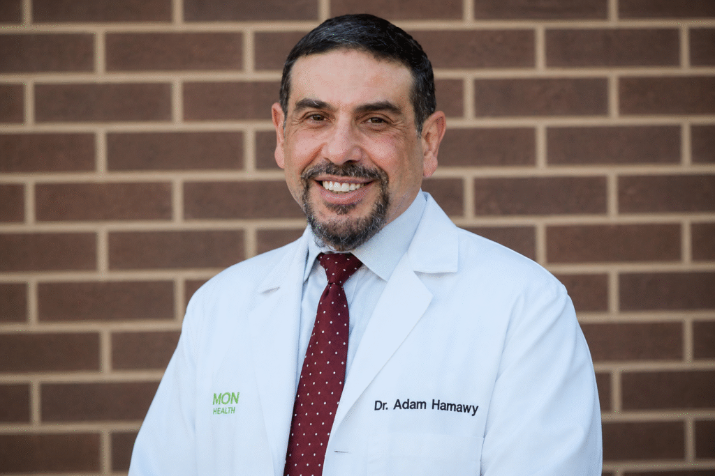 Portrait of a middle-aged man wearing a white medical coat and red tie, smiling in front of a brick wall background.
