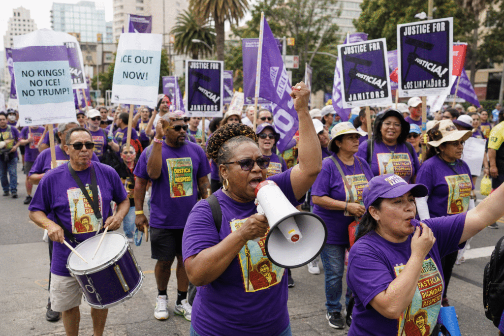 A large, diverse group of protesters marching down a city street, many wearing purple shirts and holding signs and flags, with a woman at the front speaking through a megaphone while others drum and raise their fists.