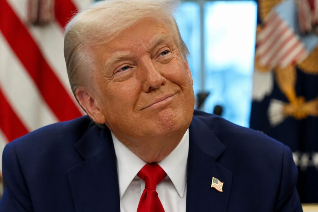 Close-up of Donald Trump in a suit and red tie, looking slightly upward with a thoughtful expression and faint smile in an office setting
