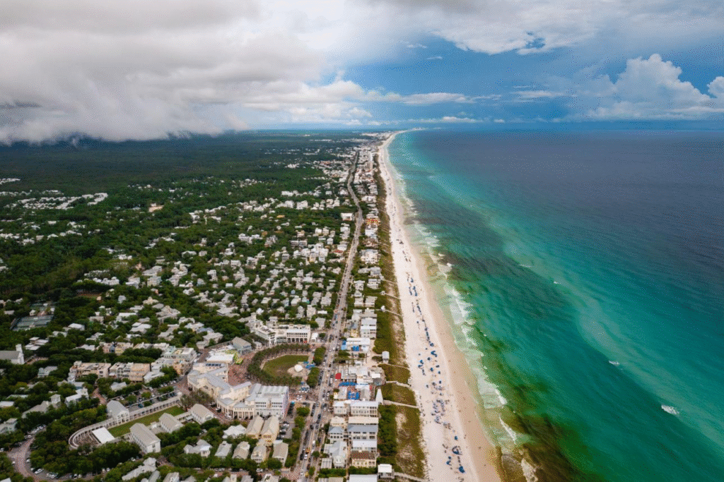 Aerial view of a Florida Gulf Coast beach showing turquoise ocean water, sandy shoreline, and a coastal town with homes, roads, and buildings stretching along the coastline under a partly cloudy sky.