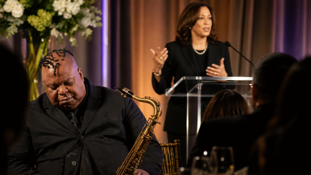 Saxophonist in a black suit appears asleep while holding his instrument at a formal awards event as a speaker talks at a podium in the background